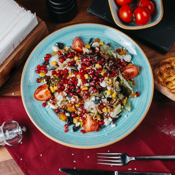 A vibrant Mediterranean salad with pomegranate, feta, corn, and tomatoes on a blue plate.