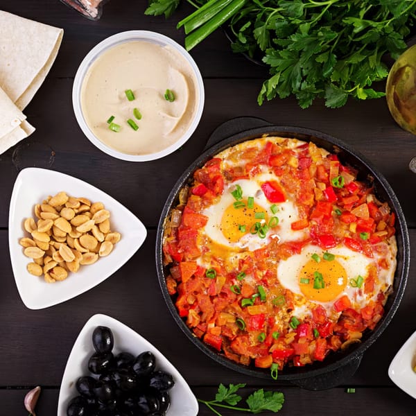 Top-down view of a Mediterranean shakshuka with eggs, tomatoes, and peppers, surrounded by hummus, pita, olives, and peanuts.