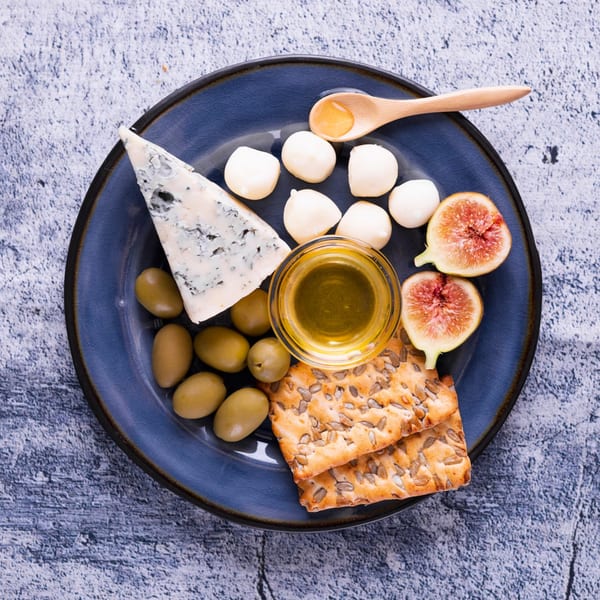 A blue plate with blue cheese, mozzarella balls, olives, figs, honey, olive oil, and crackers.